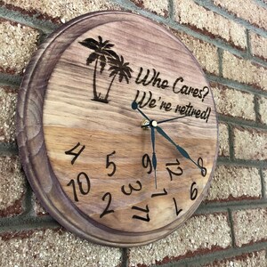 Op de afbeelding: Een houten klok met een bruine vlek. De wijzerplaat van de klok heeft zwarte Romeinse cijfers en zwarte wijzers. De klok is versierd met een in hout gebrand beeld van palmbomen en de tekst "Who Cares? We're retired!"