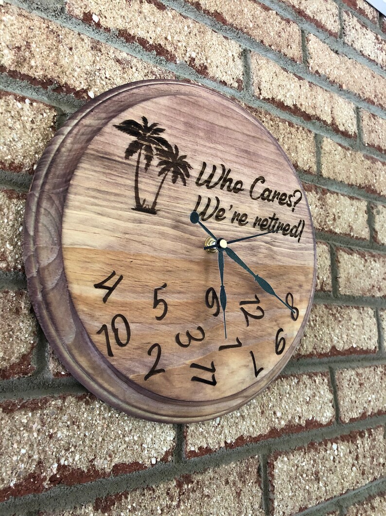 Op de afbeelding: Een houten klok met een bruine vlek. De wijzerplaat van de klok heeft zwarte Romeinse cijfers en zwarte wijzers. De klok is versierd met een in hout gebrand beeld van palmbomen en de tekst "Who Cares? We're retired!"