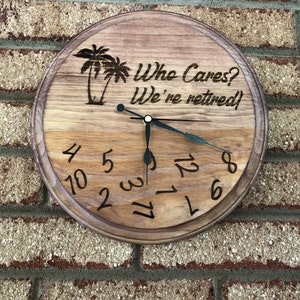 Op de afbeelding: Een houten klok met een bruine houtnerf afwerking. De wijzerplaat van de klok heeft zwarte cijfers en wijzers. De klok is versierd met een palmboom ontwerp en de tekst "Who Cares? We're retired!"
