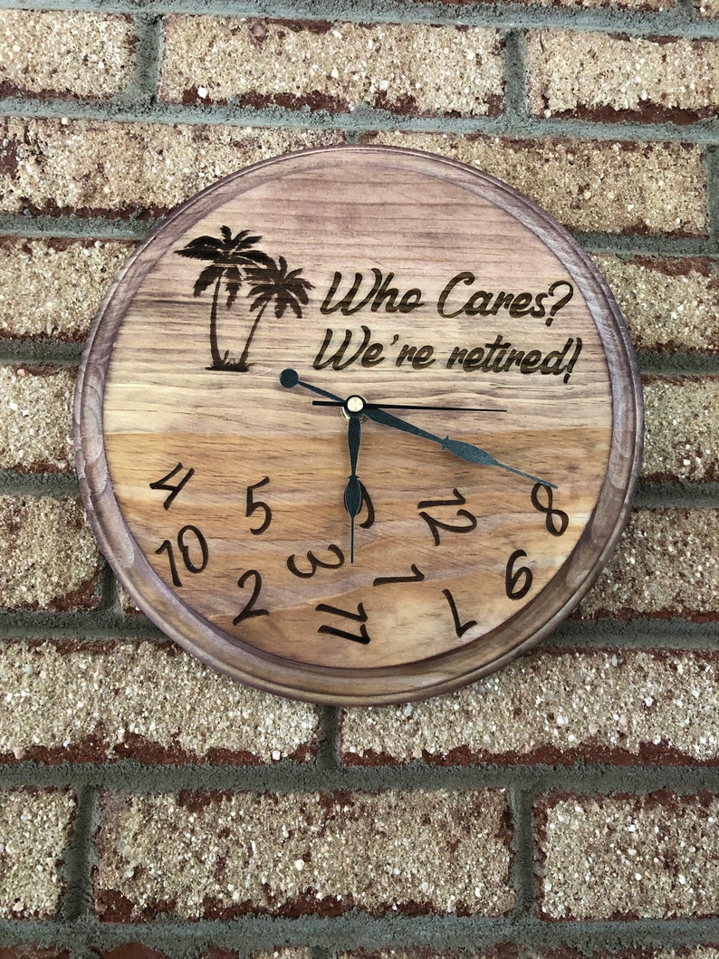 Op de afbeelding: Een houten klok met een bruine houtnerf afwerking. De wijzerplaat van de klok heeft zwarte cijfers en wijzers. De klok is versierd met een palmboom ontwerp en de tekst "Who Cares? We're retired!"