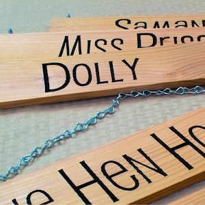 May include: Four wooden signs with black lettering hang from a silver chain. The signs read "Samantha", "Miss Drico", "DOLLY", and "The Hen House".