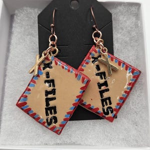 May include: A pair of dangle earrings featuring a tan, red, and blue design with the text "X-FILES". The earrings have a gold-colored X charm and are suspended from copper-colored hooks. The text "top secret folders sensitive ears" is above the earrings.