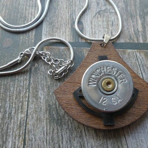Könnte beinhalten: Eine einzigartige Anhänger Halskette mit einer Winchester 12 GA Schrotpatrone auf einem Holzsockel. Der Anhänger hängt an einer silberfarbenen Kette. Das Gehäuse ist silberfarben mit goldenen Akzenten und dem Text "WINCHESTER 12 GA".