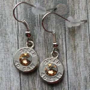 May include: A pair of silver-toned drop earrings featuring circular metal casings stamped with the text SPEER 40 S&W. Each earring is centered with a single round, faceted amber-colored crystal and includes a silver-toned hook with a clear plastic back.