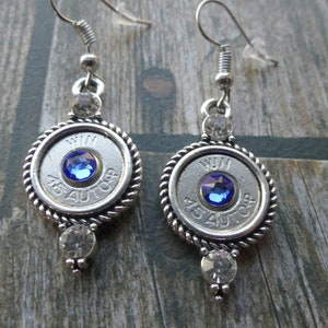 May include: A pair of silver earrings. Each earring has a circular pendant with the text "WIN 45 AUTO" and a blue gemstone. Clear crystals are set above and below each pendant. The earrings are suspended from silver-coloured ear wires.