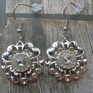May include: Silver-toned dangle earrings. Each earring features a decorative filigree design surrounding a bullet casing with a central clear crystal. The casing is marked with the text "SPEER 40 S&W".