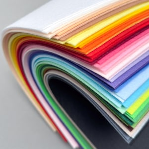May include: A collection of felt sheets in a vibrant array of colours. The sheets are arranged in a curved stack, displaying a spectrum from white to black. The colours include yellow, orange, red, pink, purple, blue, and green. The felt has a soft texture.