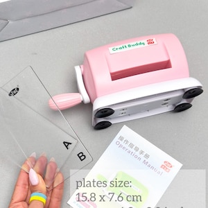 May include: A pink Craft Buddy die-cutting machine with a white base and suction cups. Two clear plastic plates are next to the machine. The plates measure approximately 15.8 x 7.6 cm. A manual is also included.