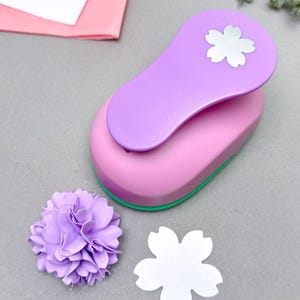 May include: A pink and purple flower-shaped paper punch with a matching paper flower and cut-out. The punch has a purple handle and a pink base with a green trim. The cut-out is white.