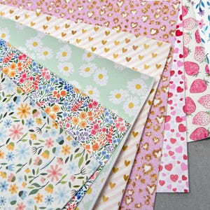 May include: A collection of patterned faux leather sheets. Designs include strawberries, hearts, gold hearts on stripes, leopard print, and floral patterns with daisies and wildflowers. The color palette is primarily pink, white, and green.