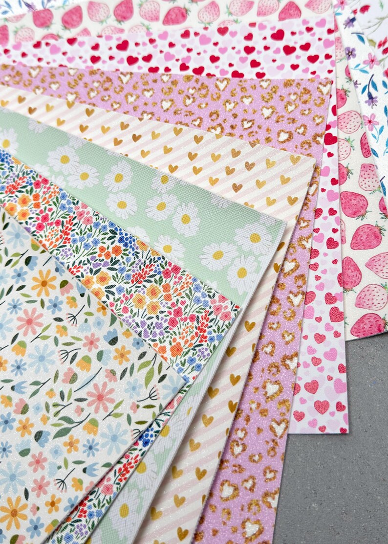 May include: A collection of patterned faux leather sheets. Designs include strawberries, hearts, gold hearts on stripes, leopard print, and floral patterns with daisies and wildflowers. The color palette is primarily pink, white, and green.