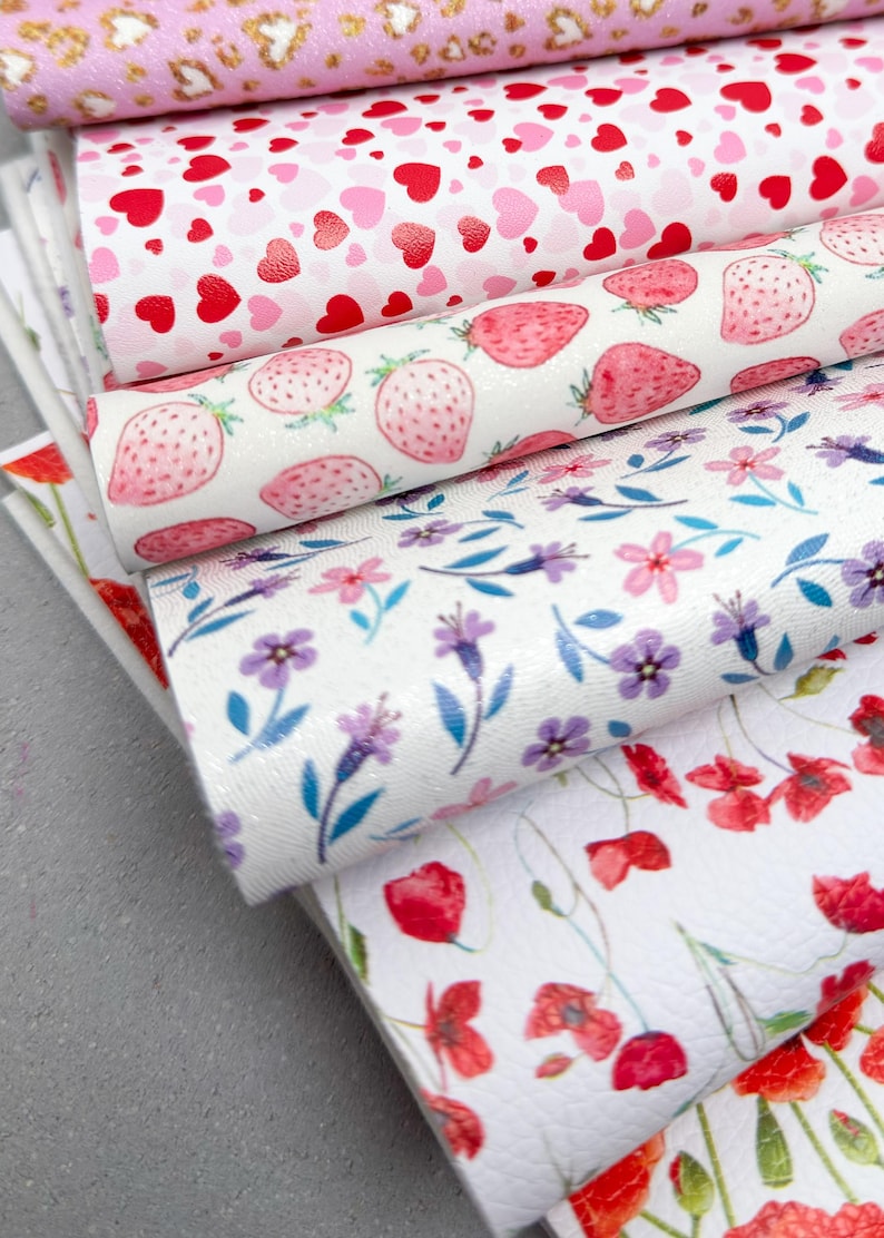 May include: A stack of decorative faux leather sheets with various patterns. Designs include pink leopard print, red and pink hearts, pink strawberries, purple flowers, and red poppies. The sheets are stacked on a gray surface.