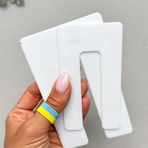 Könnte beinhalten: Zwei weiße, U-förmige Acrylrohlinge mit glatten, abgerundeten Kanten. Diese flachen, rechteckigen Komponenten verfügen über einen mittigen Ausschnitt, der eine deutliche U-Form erzeugt. Das Material ist undurchsichtig weiß und gleichmäßig.