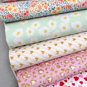May include: A stack of patterned faux leather sheets. Designs include floral, daisy, heart, and leopard prints in various colors like white, pink, and green. The sheets are rolled up, showcasing the variety of patterns available for crafting projects.