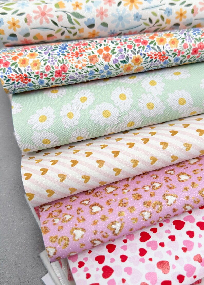 May include: A stack of patterned faux leather sheets. Designs include floral, daisy, heart, and leopard prints in various colors like white, pink, and green. The sheets are rolled up, showcasing the variety of patterns available for crafting projects.