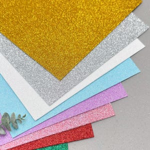 Può includere: Una pila di fogli rettangolari di cartoncino glitterato in vari colori tra cui oro, argento, bianco, azzurro, viola, rosa, rosso, verde e nero, disposti a ventaglio con un piccolo rametto di foglie verdi visibile sul lato sinistro.