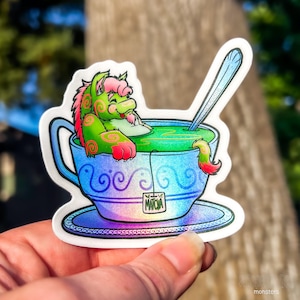 May include: A green and pink cartoon monster is relaxing in a blue and white teacup filled with green liquid. The teacup has a matching saucer and a teabag that says "MATCHA".