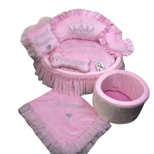 May include: Pink princess-themed dog bed set with a round bed, a pillow, a blanket, and a toy bin. The bed has a crown and the words "Princess" embroidered on it. The pillow says "Sweet Dreams". The blanket and toy bin also have the word "Princess" embroidered on them.