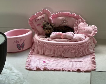 May include: A pink and white dog bed with a ruffled edge and a heart-shaped pillow that says "Coco". A small brown dog is lying in the bed. The bed also has a pink bowl that says "My Toys" and a pillow that says "Love of my life".