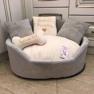 May include: A gray and white dog bed with a bone-shaped pillow and three pillows with embroidered text. The pillows say "Shh I'm sleeping", "Sweet dreams", and "Gucci". The bed itself has the text "Princess Gucci" embroidered on the side.