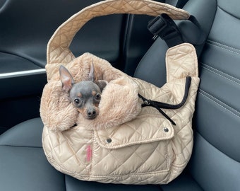 Könnte beinhalten: Ein kleiner, grau-schwarzer Hund sitzt in einer beigefarbenen, gesteppten Hundetrage mit einem Kunstfellfutter. Die Trage hat einen Reißverschluss und eine kleine Tasche auf der Vorderseite. Die Trage befindet sich auf dem Rücksitz eines Autos.