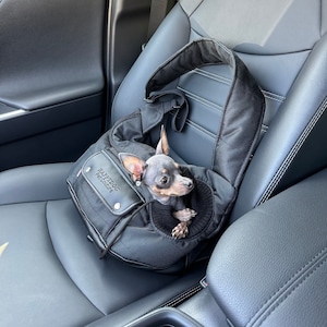 Op de afbeelding: Een zwarte dieren draagtas met de tekst "Happydog Pet Carrier" aan de voorkant. Een kleine grijze hond zit in de tas, die zich op de achterbank van een auto bevindt.