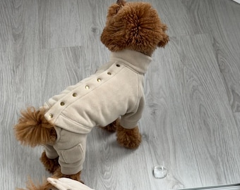 May include: A small brown dog wearing a beige fleece jumpsuit with gold button accents. The dog is standing on a light wood floor.