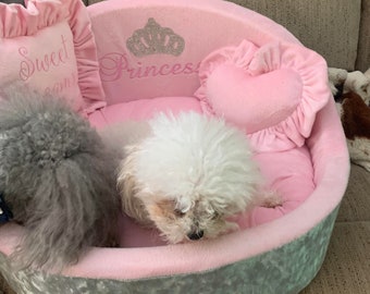 May include: A pink and gray dog bed with a heart-shaped pillow and a pillow with the text "Sweet Dreams" and "Princess" with a crown design. Two small dogs are in the bed.