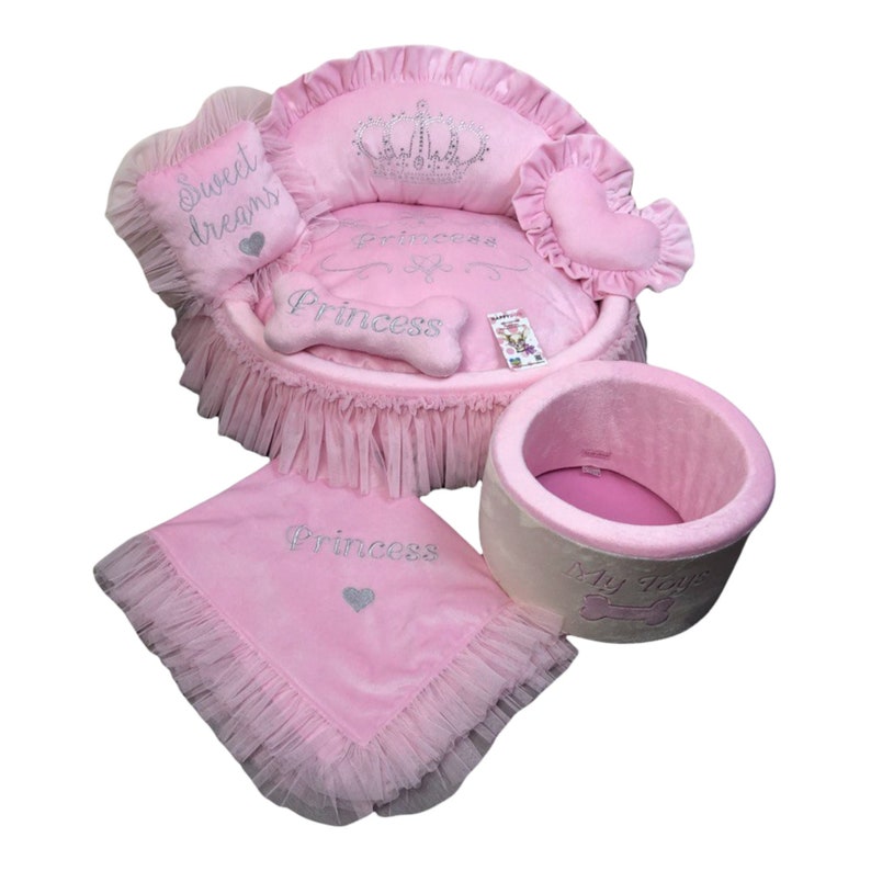 May include: Pink princess-themed dog bed set with a round bed, a pillow, a blanket, and a toy bin. The bed has a crown and the words "Princess" embroidered on it. The pillow says "Sweet Dreams". The blanket and toy bin also have the word "Princess" embroidered on them.