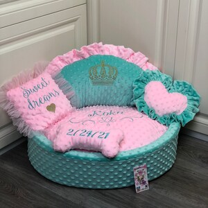 May include: A light blue and pink dog bed with a crown, heart, and bone shaped pillows. The bed is embroidered with the name "Koko" and the date "2/24/21".
