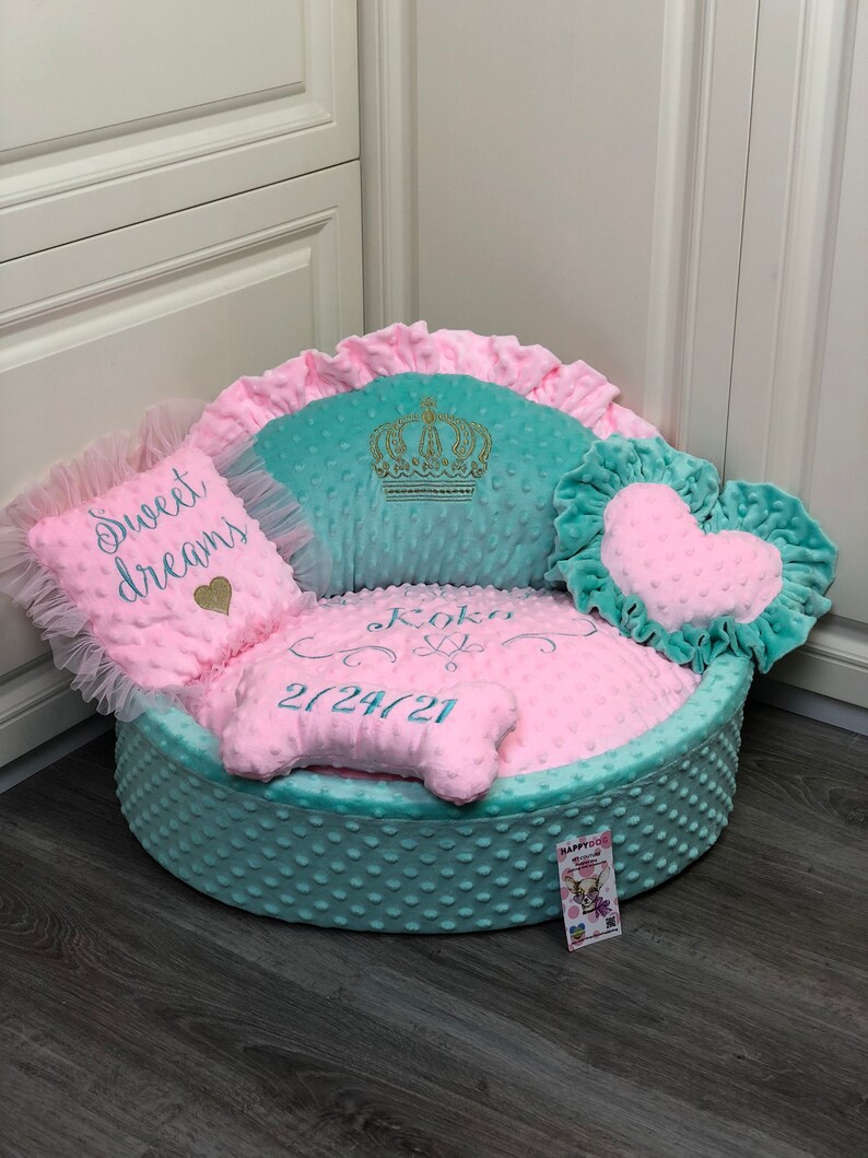 May include: A light blue and pink dog bed with a crown, heart, and bone shaped pillows. The bed is embroidered with the name "Koko" and the date "2/24/21".