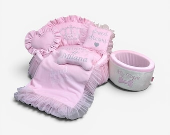 May include: A pink pet bed with ruffled tulle trim, featuring a heart-shaped pillow, a crown embroidery, and a bone-shaped pillow embroidered with the name Milana. A matching grey and pink toy bin is included, labeled with the text My Toys.