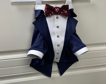 Puede incluir: Un esmoquin azul marino con camisa blanca y pajarita de lentejuelas burdeos. El esmoquin está colgado en una percha blanca.
