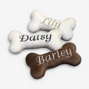 May include: Three plush bone-shaped toys made of soft fabric. Two toys are white with embroidered names in gold and black thread reading Lily and Daisy. One toy is brown with the name Barley embroidered in white thread across the center.