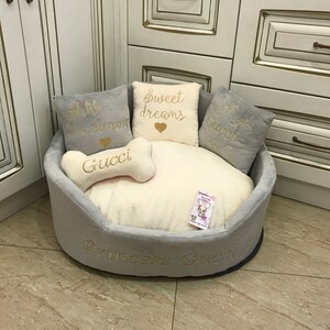 May include: A gray and white dog bed with a plush, soft interior. The bed has a bone-shaped pillow with the word "Gucci" embroidered on it. The bed also has three pillows with the words "Shh... I'm sleeping", "Sweet dreams", and "Gucci" embroidered on them. The bed is labeled "Princess Gucci" on the bottom.