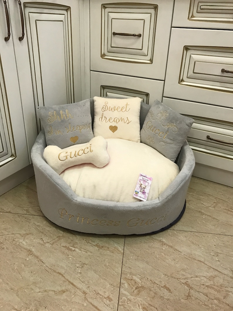 May include: A gray and white dog bed with a plush, soft interior. The bed has a bone-shaped pillow with the word "Gucci" embroidered on it. The bed also has three pillows with the words "Shh... I'm sleeping", "Sweet dreams", and "Gucci" embroidered on them. The bed is labeled "Princess Gucci" on the bottom.