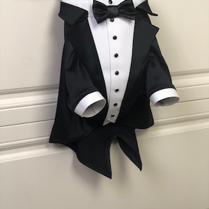 May include: A black tuxedo with a white shirt and a black bow tie. The tuxedo is hanging on a white hanger.