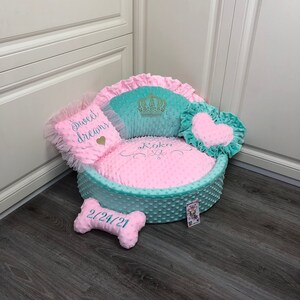 May include: A pink and teal dog bed with a crown, heart, and bone shaped pillows. The bed is embroidered with the words "Sweet Dreams" and "Koko" and the date "2/24/21".