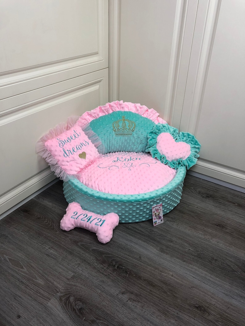 May include: A pink and teal dog bed with a crown, heart, and bone shaped pillows. The bed is embroidered with the words "Sweet Dreams" and "Koko" and the date "2/24/21".