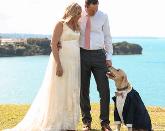Könnte beinhalten: Eine Hochzeitszene mit einem Paar und einem Hund. Die Frau trägt ein weißes Brautkleid, der Mann ein weißes Hemd und eine graue Hose. Der Hund ist in einem Smoking mit Fliege und Halsband gekleidet, vor Küstenkulisse.
