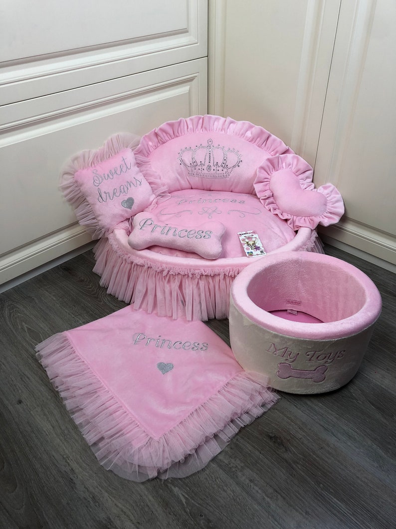 May include: Pink dog bed with a crown and the words "Princess" and "Sweet Dreams". The bed has a pink and white ruffled skirt. There is also a pink blanket with the word "Princess" and a pink and white dog toy with the words "My Toys".