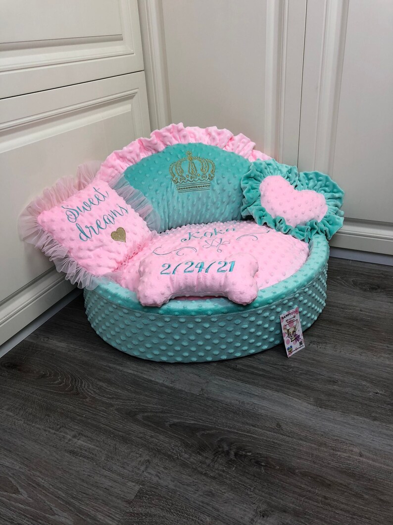 May include: A turquoise and pink dog bed with a crown, heart, and bone-shaped pillows. The bed is embroidered with the text "Koko" and "2724721".
