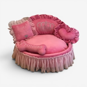 May include: A round pink pet bed with a ruffled tulle skirt, featuring a backrest with a rhinestone crown design. It includes a square pillow with the text Sweet dreams and a heart, a heart-shaped pillow, and a bone-shaped pillow labeled Princess.