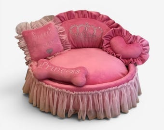 May include: A round pink pet bed with a ruffled tulle skirt, featuring a backrest with a rhinestone crown design. It includes a square pillow with the text Sweet dreams and a heart, a heart-shaped pillow, and a bone-shaped pillow labeled Princess.