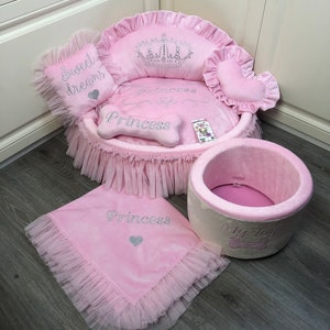May include: Pink princess dog bed with a crown, bone, and heart-shaped pillows. The bed has a ruffled skirt and a matching blanket. The bed also has a pink and white round bed with the text "My Toys" on it.
