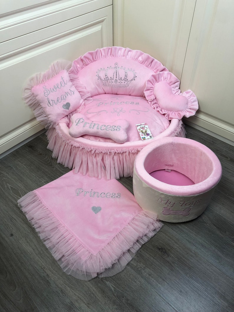 May include: Pink princess dog bed with a crown, bone, and heart-shaped pillows. The bed has a ruffled skirt and a matching blanket. The bed also has a pink and white round bed with the text "My Toys" on it.