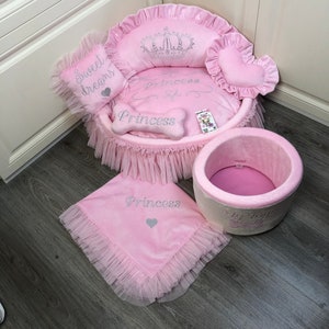 May include: Pink dog bed with a crown and the word "Princess" embroidered on it. The bed has a ruffled edge and comes with a matching pink blanket and a pink and white storage bin with the words "My Toys" and a bone.