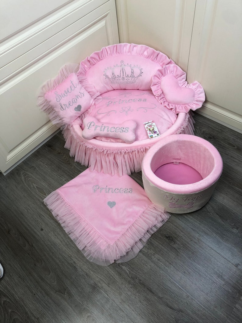 May include: Pink dog bed with a crown and the word "Princess" embroidered on it. The bed has a ruffled edge and comes with a matching pink blanket and a pink and white storage bin with the words "My Toys" and a bone.