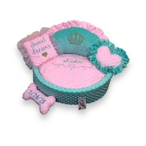 May include: A pink and teal dog bed with a crown, heart, and bone shaped pillows. The bed is embroidered with the words "Sweet Dreams" and "Koko".