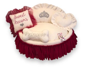 May include: A white dog bed with a red tulle skirt. The bed has a heart-shaped pillow, a bone-shaped pillow with the word "Princess" embroidered on it, and a pillow with the words "Sweet dreams" embroidered on it. The bed also has a crown-shaped pillow with rhinestones.